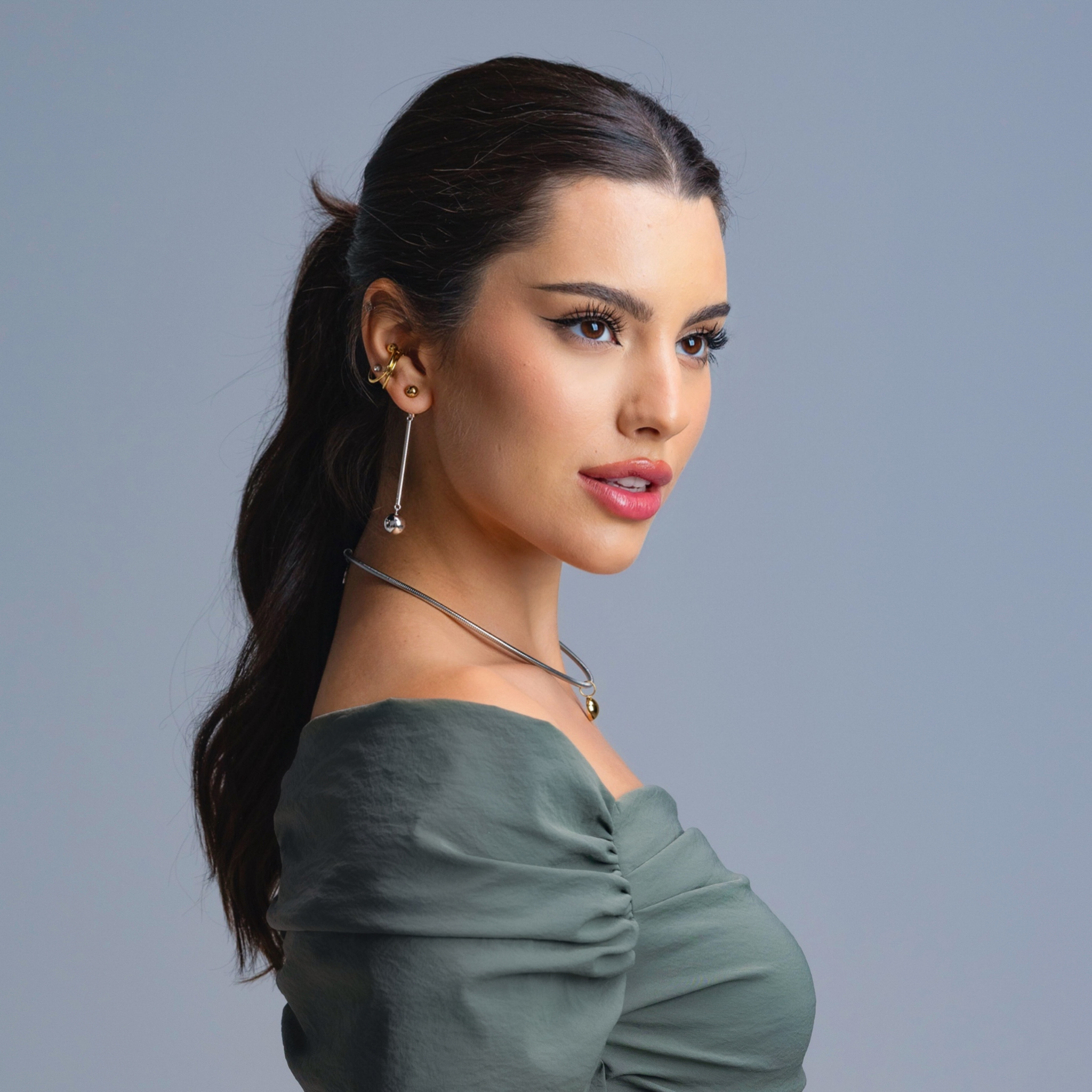 Woman wearing stacked gold ear cuffs paired with long silver drop earrings and a silver choker, styled in an off-shoulder green top against a grey background.