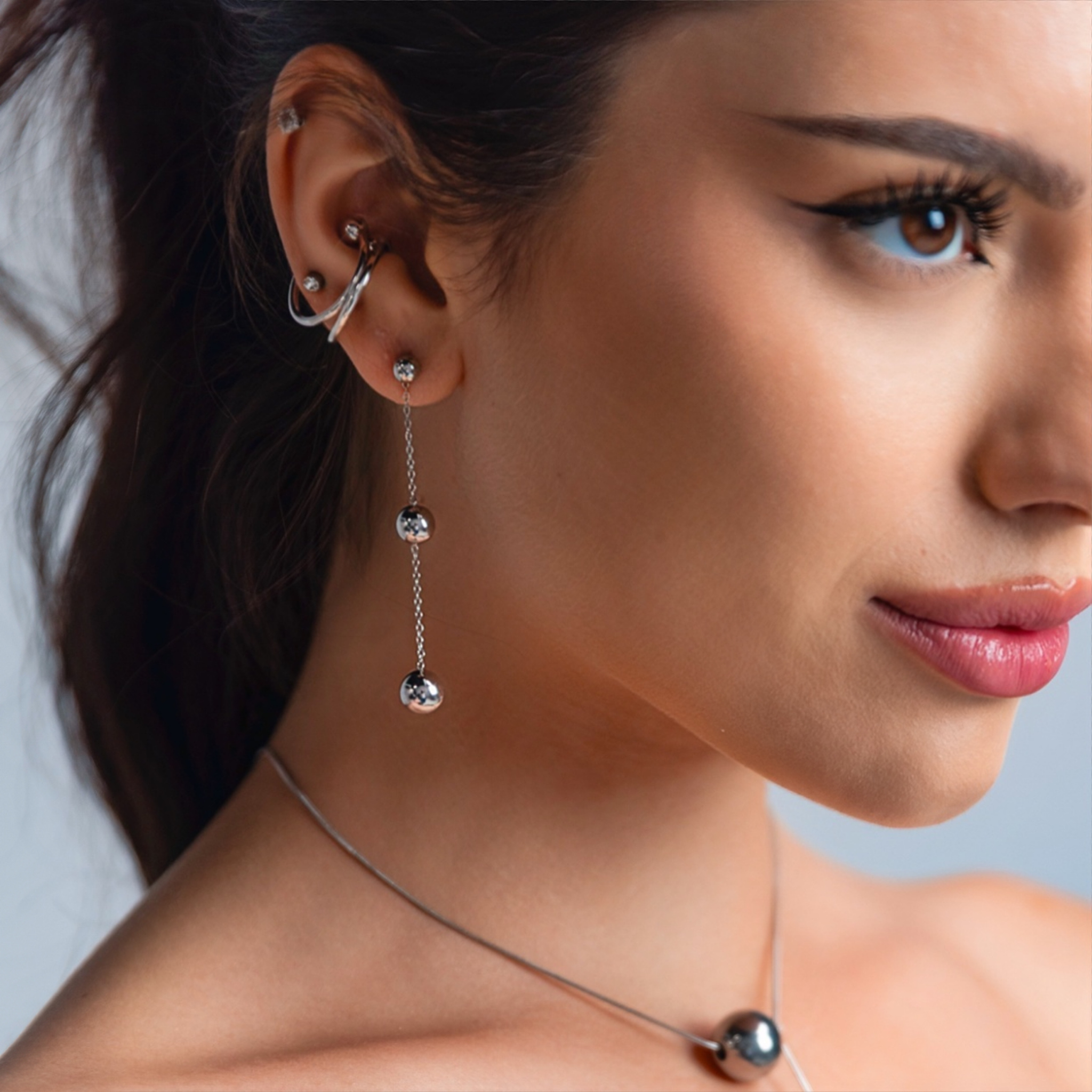 Close-up of a woman wearing silver chain drop earrings with three polished spheres, paired with stacked silver ear cuffs and a matching silver necklace.