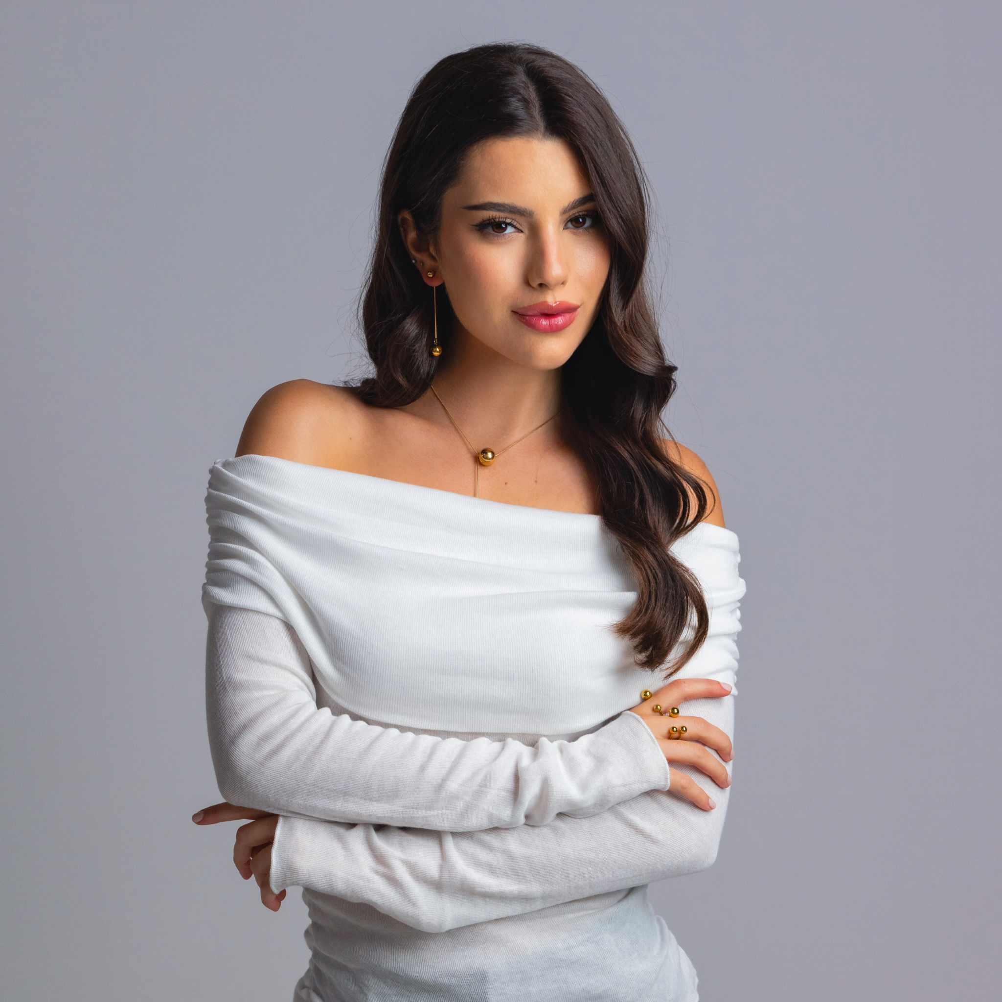 Woman wearing gold sphere earrings, a matching gold sphere necklace, and coordinating gold rings, styled in an off-shoulder white top against a grey background.