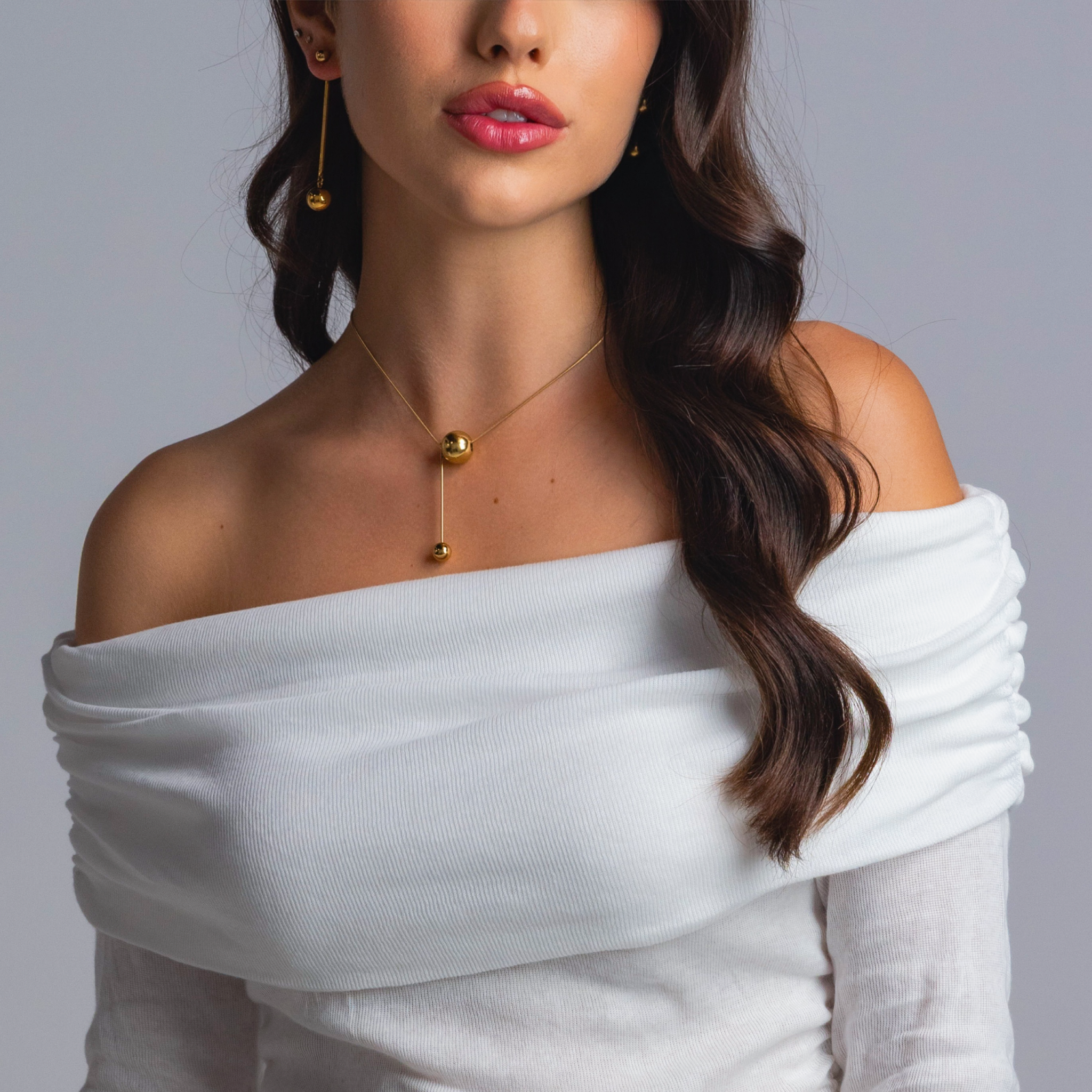 Close-up of a woman wearing gold sphere drop earrings and a matching gold sphere pendant necklace, styled with an off-shoulder white top.