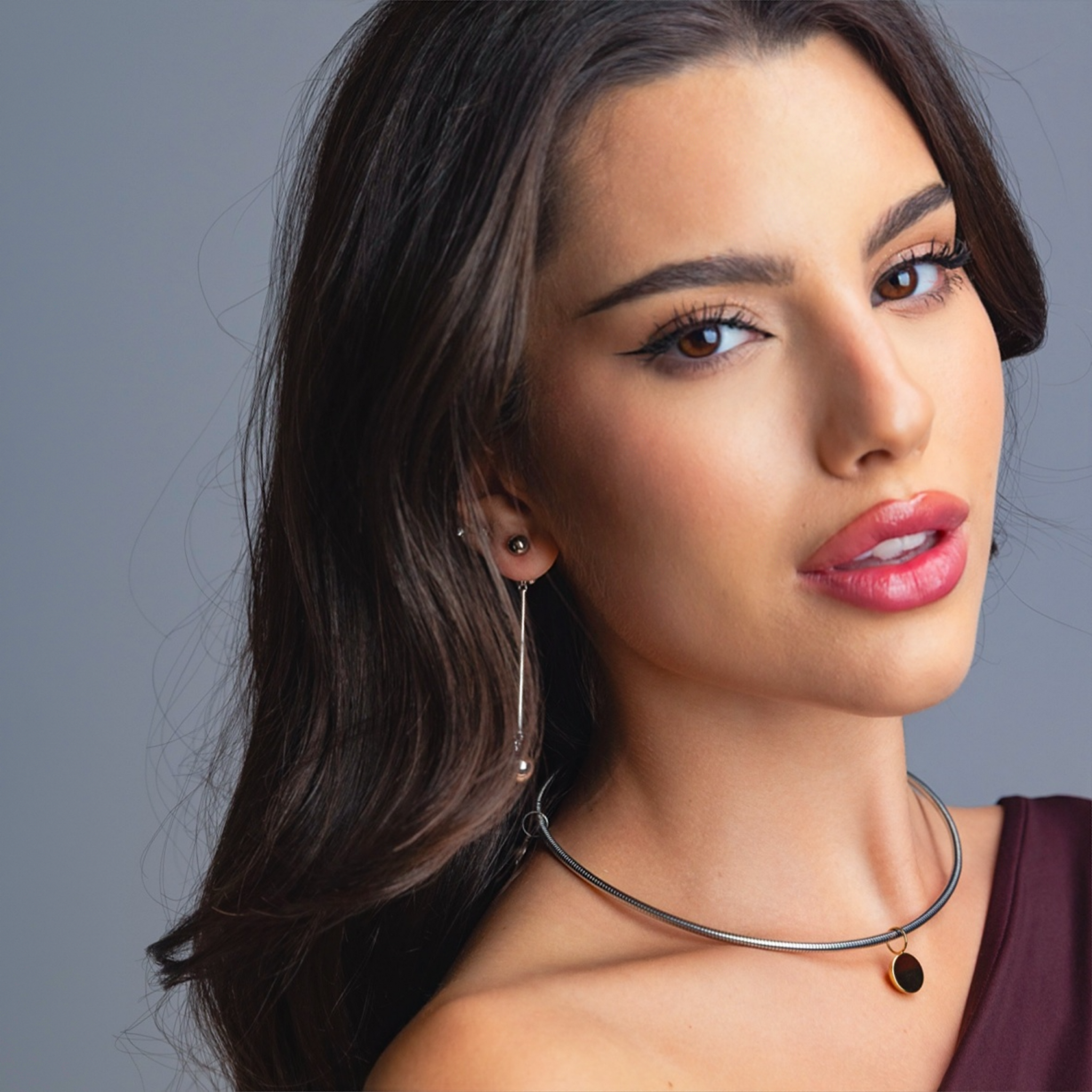 Woman wearing long silver drop earrings and a sleek silver choker necklace with a round gold pendant, styled in a purple one-shoulder top against a grey background.