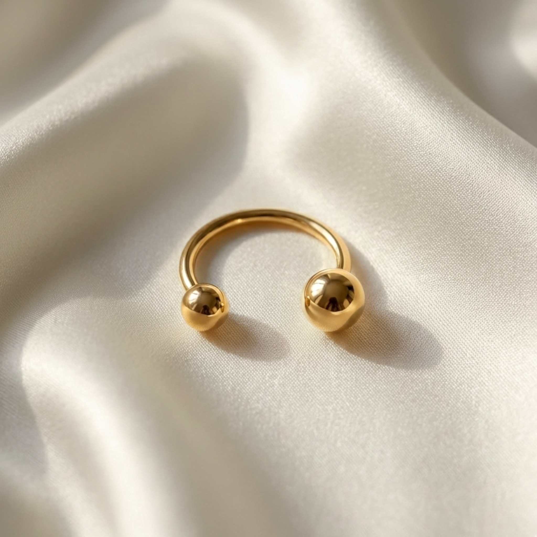 Gold open-band ring with two polished sphere ends displayed on soft ivory satin fabric.