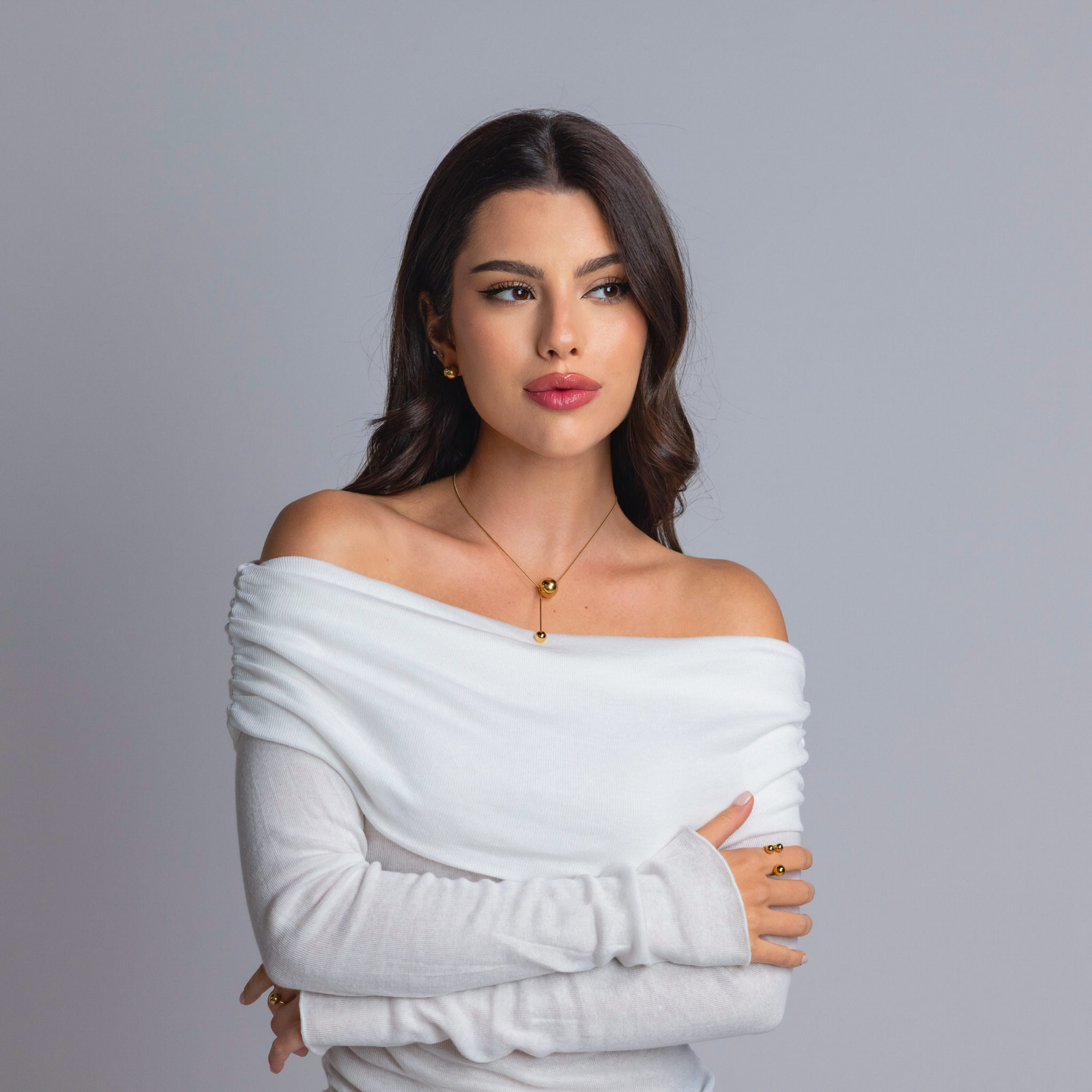 Woman wearing a gold sphere pendant necklace with matching gold earrings and rings, styled in an off-shoulder white top against a grey background.