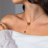 Close-up of a woman wearing a gold sphere pendant necklace with a long bar drop, styled with a matching gold ring and an off-shoulder white top.
