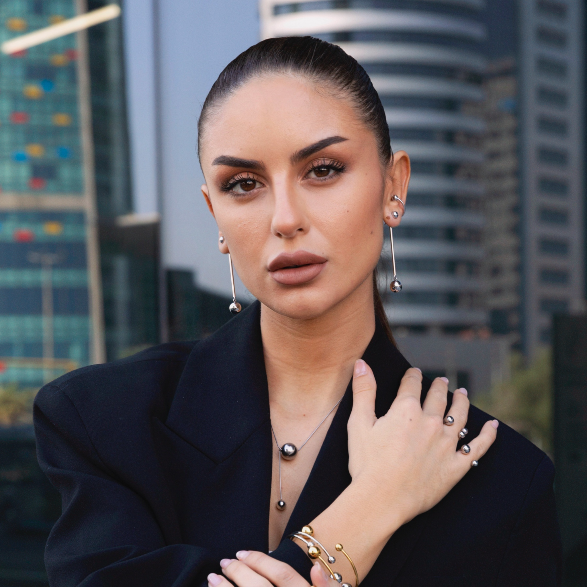 Woman wearing silver sphere drop earrings, a matching silver pendant necklace, stacked ear cuffs, and coordinating gold and silver bracelets and rings, styled in a black blazer with a cityscape background.