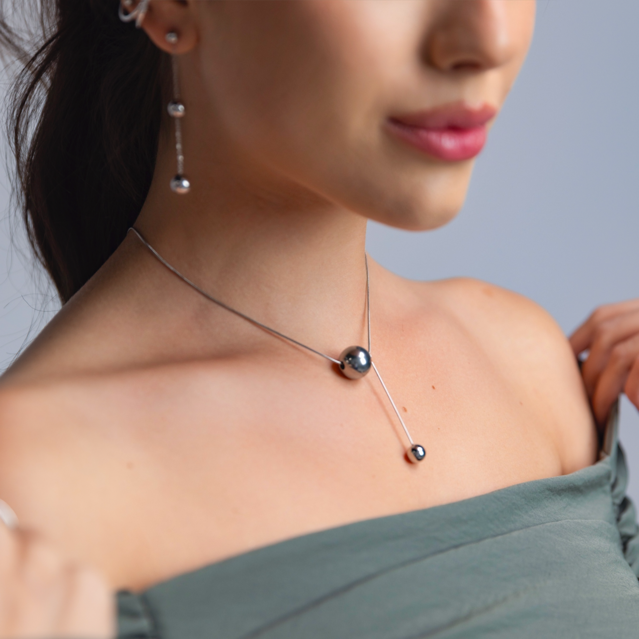Close-up of a woman wearing a silver sphere pendant necklace with a long bar drop, paired with silver chain drop earrings and stacked ear cuffs.