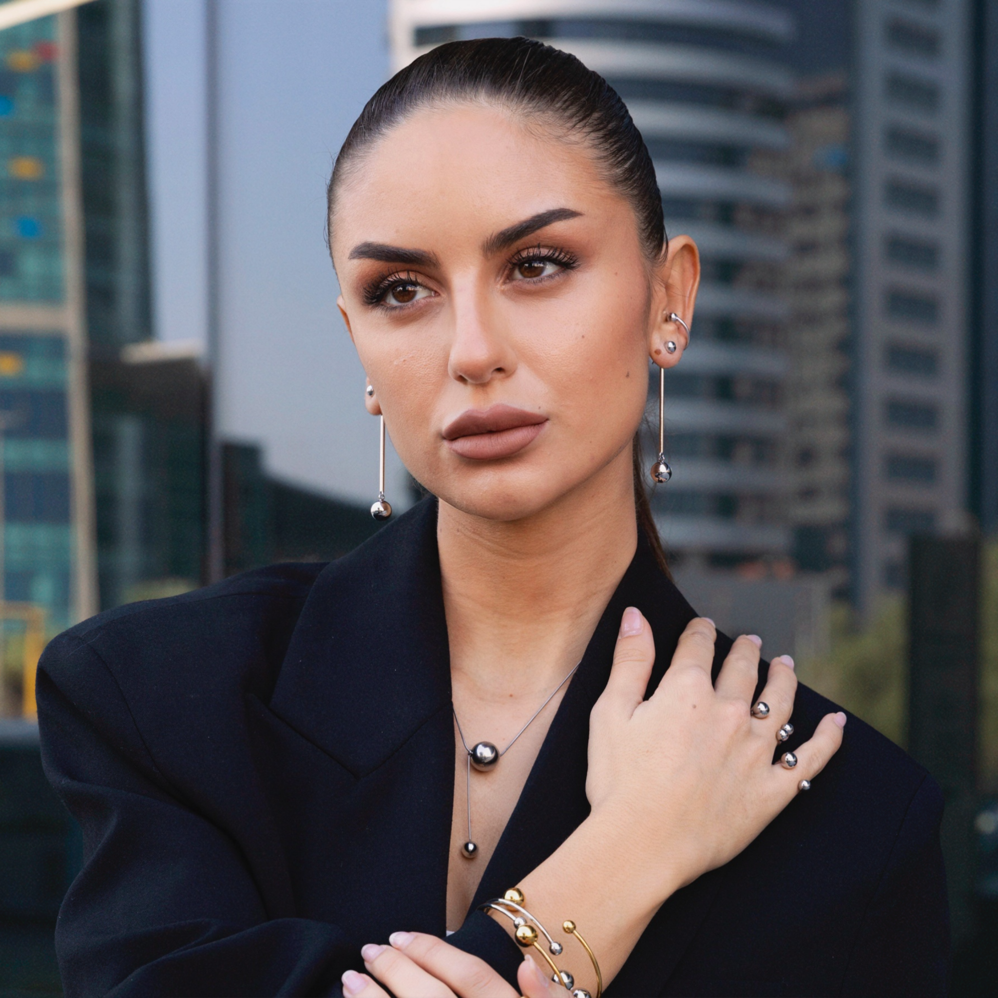 Woman wearing silver sphere drop earrings, a matching silver pendant necklace, stacked ear cuffs, and coordinating gold and silver bracelets and rings, styled in a black blazer with a cityscape background.