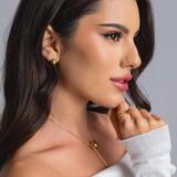 Close-up side view of a woman wearing gold sphere stud earrings, a gold sphere pendant necklace, and a gold open-band ring with polished spheres, styled in an off-shoulder white top.