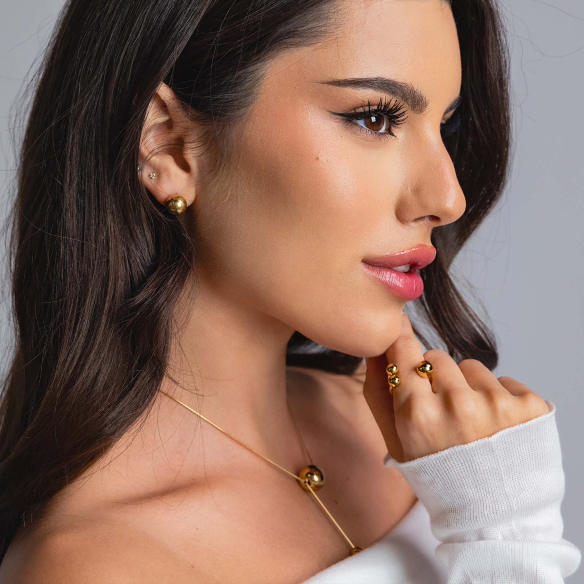 Close-up side view of a woman wearing gold sphere stud earrings, a gold sphere pendant necklace, and a gold open-band ring with polished spheres, styled in an off-shoulder white top.
