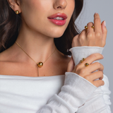 Close-up of a woman wearing a gold sphere pendant necklace, matching gold stud earrings, and two gold open-band rings with polished spheres, styled in an off-shoulder white top.
