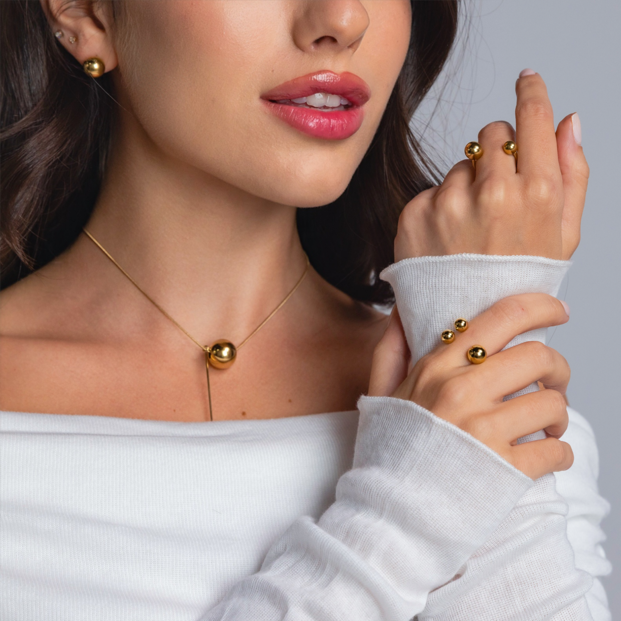 Close-up of a woman wearing a gold sphere pendant necklace, matching gold stud earrings, and two gold open-band rings with polished spheres, styled in an off-shoulder white top.