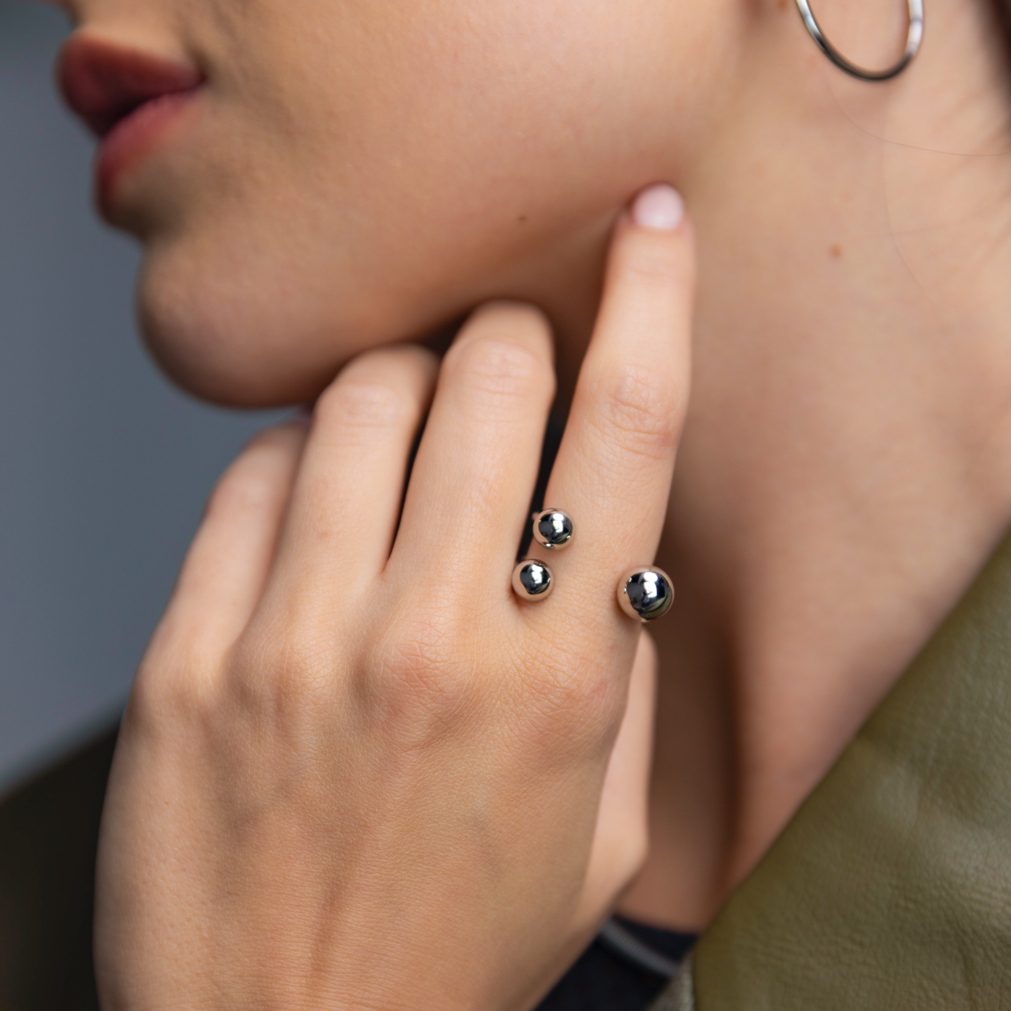 Close-up of a woman wearing a silver open-band ring with three polished spheres, shown against her cheek with a silver hoop earring visible.