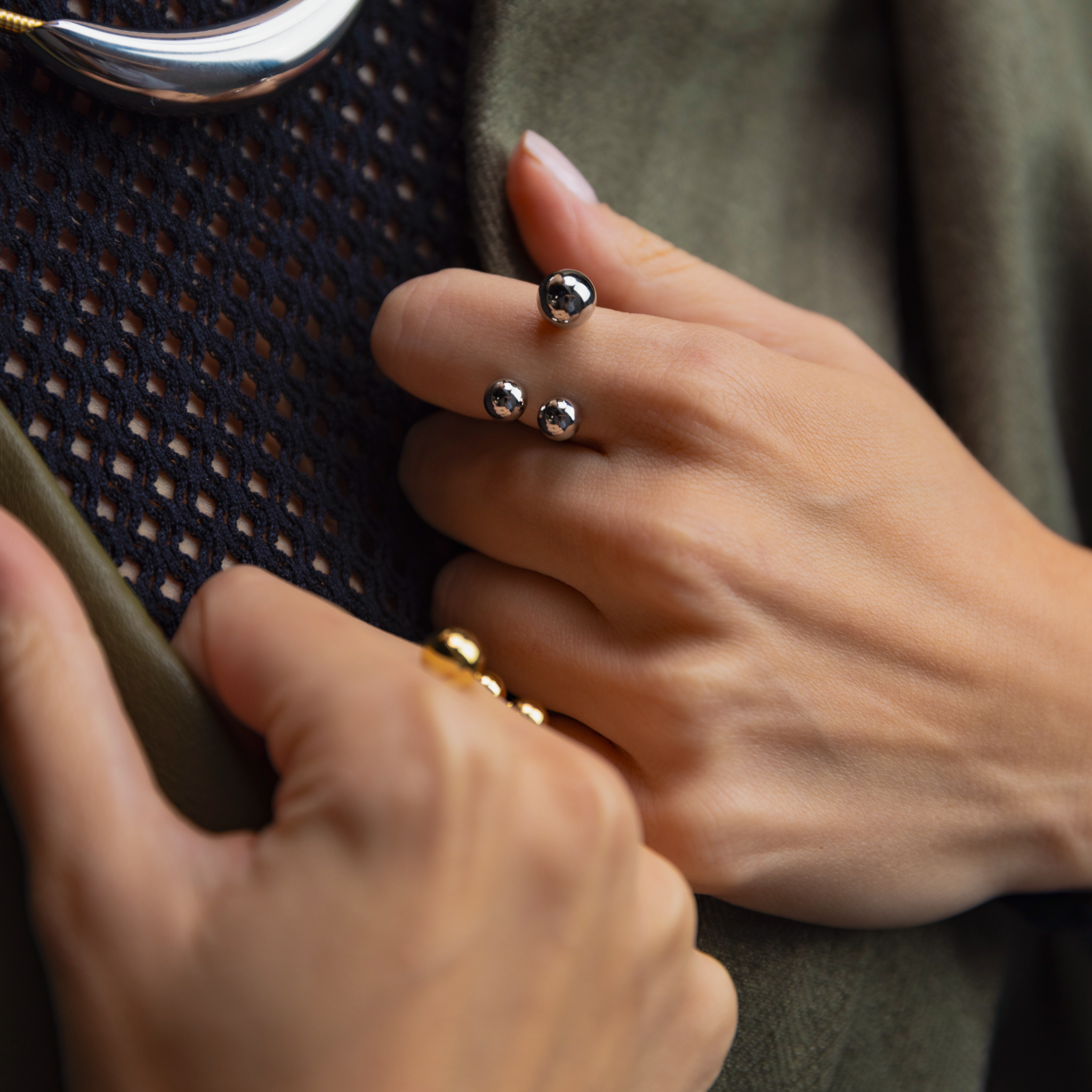 Close-up of a woman's hand wearing a silver open-band ring with three polished spheres, styled alongside a gold ring and textured black and olive green clothing.